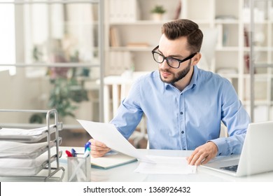 Young Serious Businessman In Eyeglasses Reading Financial Paper Or Contract While Sitting By Desk In Office