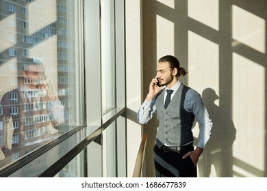 Young serious businessman looking through window on sunny day while consulting one of clients by smartphone on working day - Shutterstock ID 1686677839