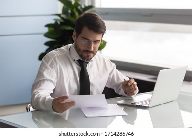 Young serious businessman reading document and analyzing statistics. Employee working on business research project, using laptop, man worker look at business letter or report. - Shutterstock ID 1714286746