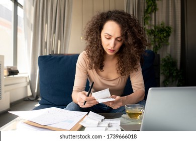 Young serious businesswoman with long wavy hair looking through payment bills while sitting on couch in front of laptop