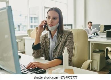 Young Serious Businesswoman Pushing Keyboard Key While Looking At Computer Screen And Talking To Someone On Smartphone At Work