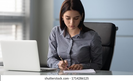 Young Serious Businesswoman Sign Up Document At Work. Tired Woman Employee Writing Report On Paper Near Modern Laptop. Success Business Deal Concept.