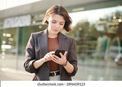 Joven empresaria seria en ropa informal inteligente recorriendo contactos en smartphone mientras iba a llamar por teléfono en un ambiente urbano