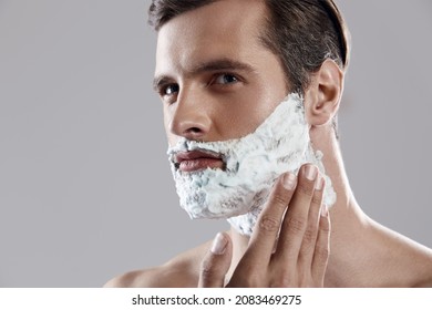 Young Serious Caucasian Man Applying Shaving Foam On His Face. Close Up Of Face Of Dark-haired Handsome Bearded Guy. Concept Of Face Care. Idea Of Male Beauty. Isolated On White Background In Studio