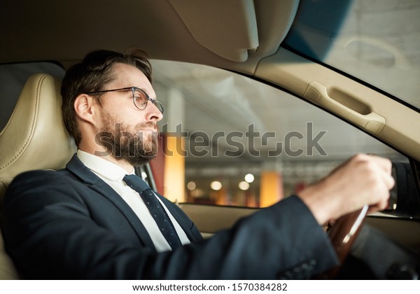 Young serious driver in eyeglasses and in\
suit holding the wheel and driving the\
car