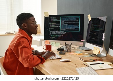 Young serious IT engineer in smart orange shirt looking at coded data on computer screens while typing on keyboard by workplace