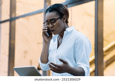 Young serious executive woman worried with problem discussing working process by smartphone while checking information in tablet near office building - Powered by Shutterstock