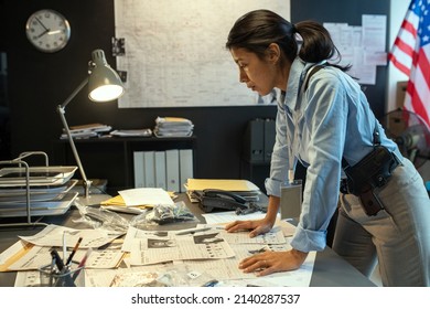 Young serious female detective in uniform bending over workplace with documents while learning criminal profiles during investigation - Shutterstock ID 2140287537