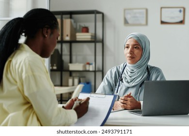 Young serious female doctor in hijab looking at her patient signing medical document or agreement about long term treatment after consultation - Powered by Shutterstock