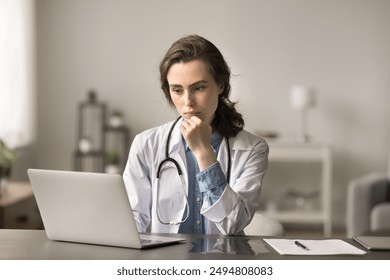 Young serious focused doctor, GP or intern studying on laptop, accessing electronic Health records, review patient medical history, analyzing data, make decisions about diagnosis and treatment plan - Powered by Shutterstock