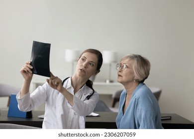 Young serious focused female therapist in white coat hold roentgen film, show x-ray with result of radiology to elderly patient, explain diagnosis of trauma and shot of bones, tells about treatment - Powered by Shutterstock