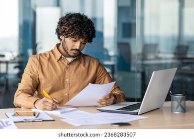Young serious focused and thinking businessman paper work, successful hispanic man reviewing contract reports and invoices, sitting at desk inside office with laptop, checking and filling form.
