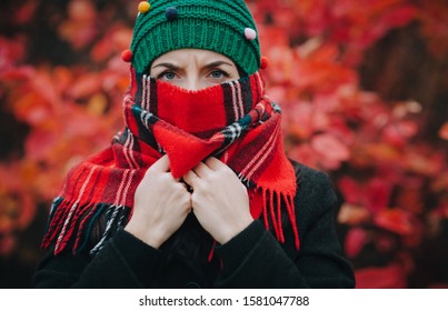 A Young Serious Girl With Her Eyebrows Shifted In A Green Knitted Hat And A Scarf Covering Her Face Is Standing Against The Backdrop Of Autumn Leaves. The Concept Of Anger,  Anonymity, Cold And Wind.