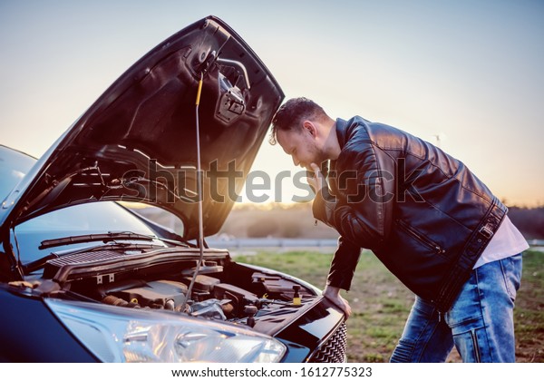 Young serious handsome caucasian bearded blond man\
in leather jacket looking engine of his car under the hood.\
Something went wrong.