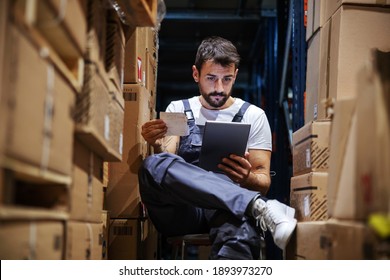 Young Serious Hardworking Blue Collar Worker In Overalls Sitting On Chair In Storage Surrounded By Boxes And Checking On Delivery On Tablet. Import And Export Firm Interior.