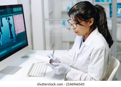 Young Serious Hispanic Female Scientist Making Notes In Her Notepad While Sitting By Workplace In Front Of Computer During Research