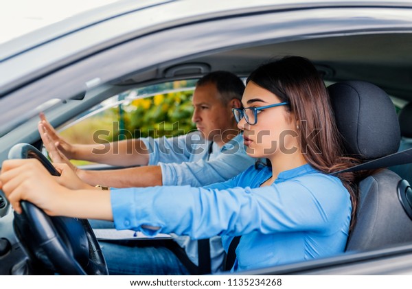  Young serious\
inexperienced woman driving a car in alert, worried instructor man\
sitting aside and looking nervous at the road, dangerous situation.\
Failed Driving test.