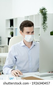 Young Serious Male Designer With Protective Mask On Face Looking At Computer Screen While Networking By Desk In Office Environment