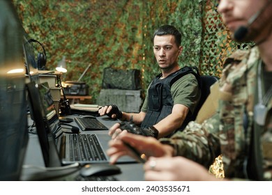 Young serious male military officer in protective body armor sitting by workplace in front of computer screen next to colleague