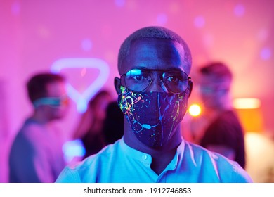 Young Serious Man Of African Ethnicity In Eyeglasses And Protective Mask Looking At You While Standing Against His Friends Enjoying Home Party