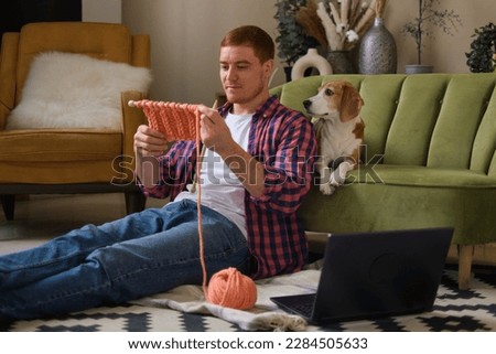 Young serious man looking at laptop. Man learning new hobby, knitting on needles. Knitting project in progress.
