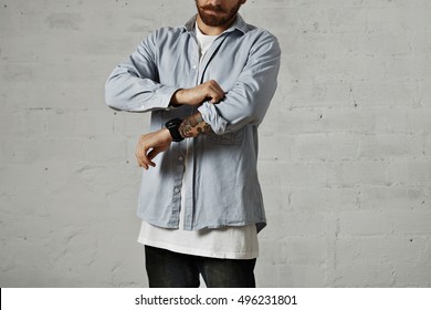 Young Serious Man Wearing A White T-shirt And A Light Blue Denim Shirt Rolling Up His Right Sleeve Showing A Tattooed Arm Isolated On White.