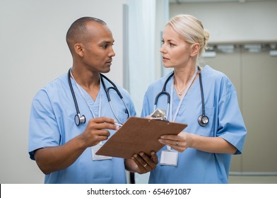 Young Serious Nurses Discussing About Medical Report. Young African Doctor Checking Patient Details Before Entering Operation Theatre. Male And Female Nurse Working Together At Medical Clinic.