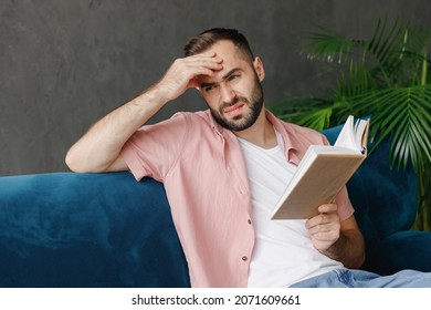 Young Serious Pensive Minded Sad Depressed Caucasian Man Wear Casual Clothes Reading Book Novel Prop Up Forehead Look Aside Sitting On Blue Sofa At Home Flat Indoors Rest Relax On Weekends Free Time.