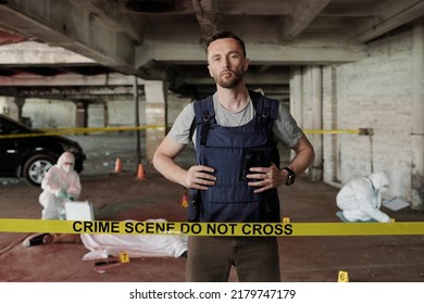 Young serious policeman in bulletproof vest standing behind crime scene tape and looking at camera against forsenic experts working