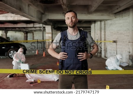 Young serious policeman in bulletproof vest standing behind crime scene tape and looking at camera against forsenic experts working
