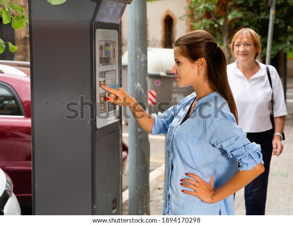 Young serious positive woman paying for\
parking in modern parking meter on city\
street