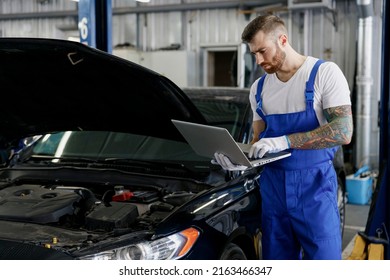 Young serious professional technician car mechanic man in blue overalls t-shirt use hold laptop pc computer make diagnostics fix problem with raised hood work in vehicle repair shop workshop indoor. - Powered by Shutterstock