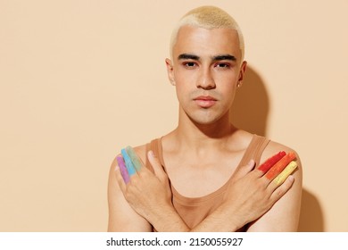 Young Serious Sexy Blond Latin Gay Man 20s With Make Up Fingers Painted In Rainbow Flag Colors Wear Beige Tank Shirt Hold Hands Crossed Folded Isolated On Plain Light Ocher Background Studio Portrait.