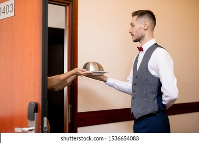 Young Serious Waiter Passing Cloche With Food To One Of Hotel Guests While Standing By Open Door