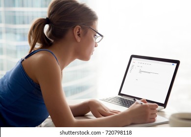 Young Serious Woman In Glasses Looks At Laptop Screen, Writing Notes. Girl Composing Email Reply To Potential Employer, University Admissions. Corporate Correspondence Or School Communication Concept.