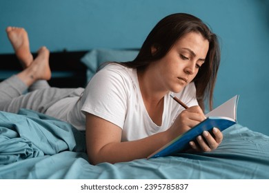 Young serious woman laying on bed and writing a diary. Caucasian lady with notebook and pen in her hands. Bright home interior in the morning. Copy space - Powered by Shutterstock