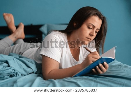 Young serious woman laying on bed and writing a diary. Caucasian lady with notebook and pen in her hands. Bright home interior in the morning. Copy space