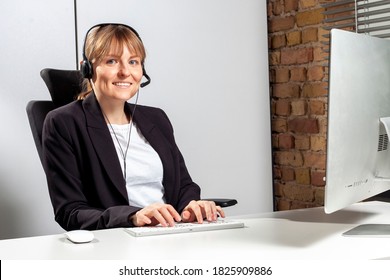 Young Service Employee Sits Smiling In Front Of The Monitor With A Headset And Consults Customers By Phone Who Need Help