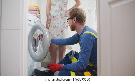 Young Service Man In Uniform Installing New Washing Machine And Explaining Woman How To Use It Sitting On Floor In Bathroom Or Laundry Room