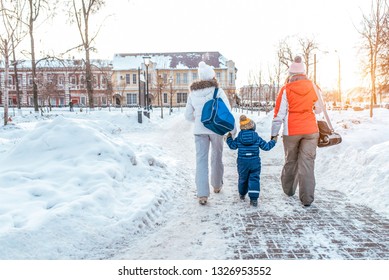 A Young, Seven Woman Mom With Son 4-5 Years Old And Girlfriend Returning Home After A Sporting Event. In Hands Of Bag With Clothes. View From Rear On Background Of Snowdrifts And Streets In City.