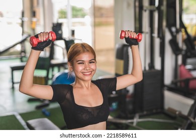 A Young And Sexy Asian Woman Smiles While Doing Seated Dumbbell Shoulder Presses On A Flat Bench At The Gym. Wearing A Black Crop Top And Bike Shorts.