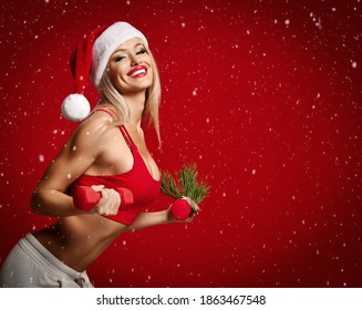 Young Sexy Blonde Fitness Woman After Diet In Sport Wear Red Sport Bra Top, Pants And Santa Hat Does Exercises Workout With Red Dumbbells Under Snow. Shape, Figure, Christmas, New Year, Celebration