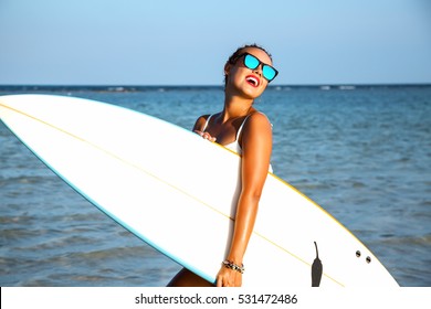 Young sexy blonde woman with long curly hairs and sportive tanned body. Smiling and  posing with surf board near blue ocean in sunny summer day. Wearing stylish mirrored sunglasses. Ready for surfing