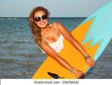 Young sexy blonde woman with long curly hairs and sportive tanned body. Smiling and posing with surf board near blue ocean in sunny day. Wearing stylish round sunglasses. Ready for surfing. Close up