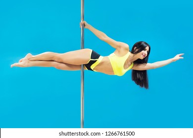 Young Sexy Brunette Making Superman Element Hanging On Pole At Blue Background. Fit Smiling Poledancer In Yellow Pole Outfit Performing Horizontal Plank. Poledancing Exciting Fitness. Copy Space