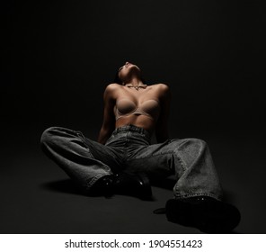 Young sexy brunette woman fitness model in pearl necklace, bra, wide jeans, and massive boots sits on floor with head thrown back over dark background. Fashion, vogue, stylish female look concept