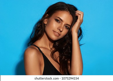 Young sexy slim tanned woman in black swimsuit posing against blue background. Closeup Fashion portrait of beautiful girl with long wavy brunette hair. Swimwear or bikini model