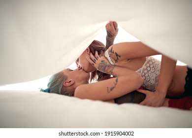 Young sexy tattooed lesbian couple in a passionate hug kissing in an intimate atmosphere under the white sheet. Love, relationship, sex, lgbt