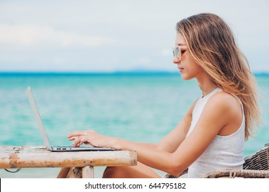 Young Sexy Woman Freelancer In Dress Sitting At The Table On Ocean Background, Using Laptop On The Beach. Girl Freelancer Working