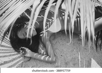 young sexy woman laying on sand beach under palm tree leaf, artistic shadow pattern on body, white bikini swim suit, tanned skin, slim body, black and white photography